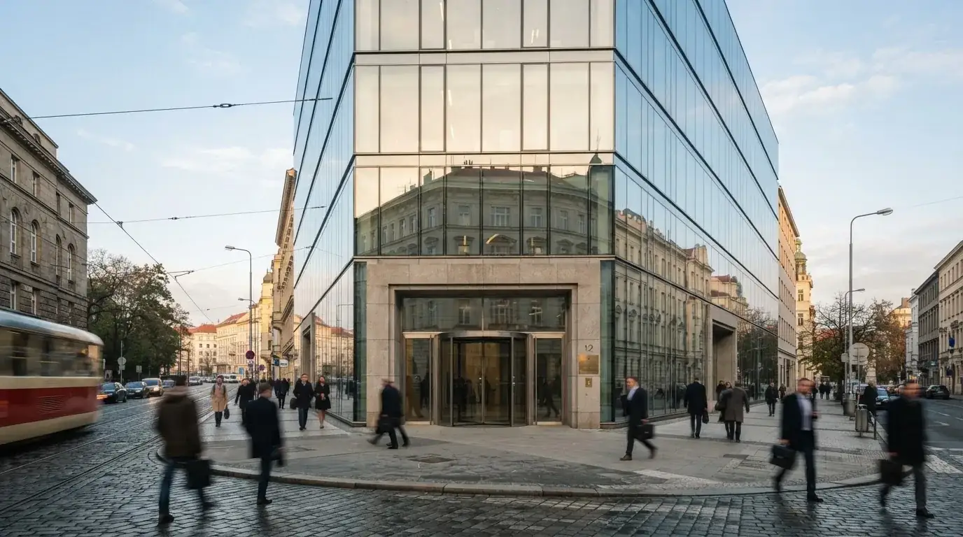Fasáda moderní administrativní budovy s skleněnými okny odráží ranní oblohu nad rušnou pražskou ulicí.
